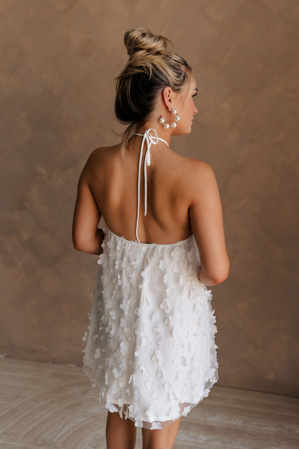 Upper body back view of female model wearing the Meredith White Embellished Floral Dress that has white sheer fabric with 3d floral embellishments, white lining, and a halter neck.
