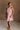 Full body front view of female model wearing the Eva Pink Sheer Tie Back Mini Dress that has pink sheer fabric with pink lining and a halter neck with a bow tie back