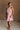 Full body front view of female model wearing the Eva Pink Sheer Tie Back Mini Dress that has pink sheer fabric with pink lining and a halter neck with a bow tie back