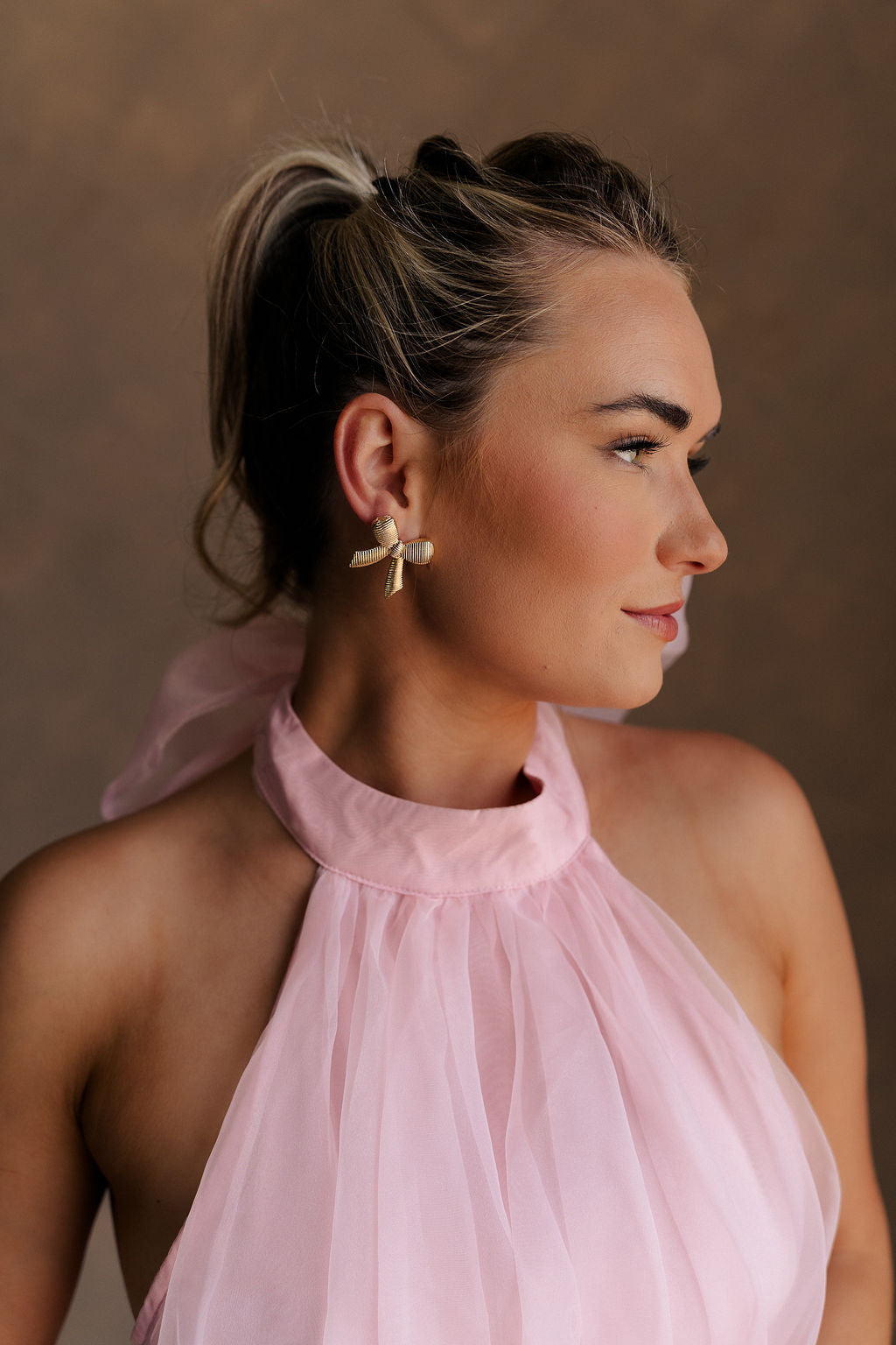 Side view of female model wearing the Lucia Gold Ribbed Bow Studs which features  gold ribbed bows with back clasp closure