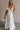 Full body front view of female model wearing the Gianna Ruffle Short Sleeve Tiered Maxi Dress in White which features Lightweight Fabric, Tiered Body, Midi Length, Pockets on each side, V-Neckline with a Tie Detail and Ruffle Short Sleeves. the dress is available in pink, aqua blue and white.