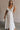Full body front view of female model wearing the Gianna Ruffle Short Sleeve Tiered Maxi Dress in White which features Lightweight Fabric, Tiered Body, Midi Length, Pockets on each side, V-Neckline with a Tie Detail and Ruffle Short Sleeves. the dress is available in pink, aqua blue and white.