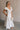 Full body side view of female model wearing the Gianna Ruffle Short Sleeve Tiered Maxi Dress in White which features Lightweight Fabric, Tiered Body, Midi Length, Pockets on each side, V-Neckline with a Tie Detail and Ruffle Short Sleeves. the dress is available in pink, aqua blue and white.