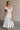 Full body side view of female model wearing the Gianna Ruffle Short Sleeve Tiered Maxi Dress in White which features Lightweight Fabric, Tiered Body, Midi Length, Pockets on each side, V-Neckline with a Tie Detail and Ruffle Short Sleeves. the dress is available in pink, aqua blue and white.