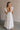 Full body back view of female model wearing the Gianna Ruffle Short Sleeve Tiered Maxi Dress in White which features Lightweight Fabric, Tiered Body, Midi Length, Pockets on each side, V-Neckline with a Tie Detail and Ruffle Short Sleeves. the dress is available in pink, aqua blue and white.