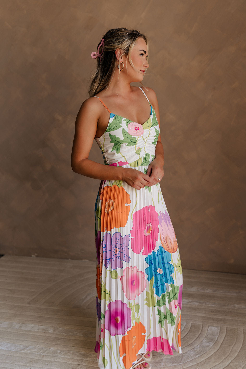 Full body side view of model wearing the Alia Pleated Floral Maxi Dress that has multi-colored fabric with a floral print, pleating, thin straps, and a sweetheart neck. Model is holding skirt.