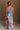 Full body back view of model wearing the Alia Pleated Floral Maxi Dress that has multi-colored fabric with a floral print, pleating, thin straps, and a sweetheart neck. Model is holding skirt.