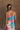 Upper body back view of model wearing the Alia Pleated Floral Maxi Dress that has multi-colored fabric with a floral print, pleating, thin straps, and a sweetheart neck. Model is holding skirt.