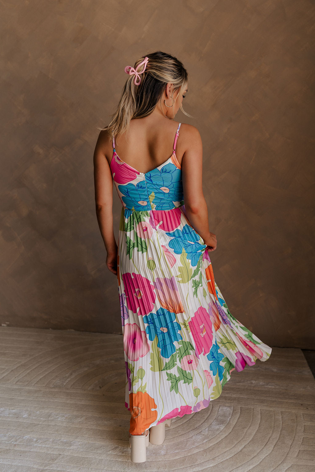 Full body back view of model wearing the Alia Pleated Floral Maxi Dress that has multi-colored fabric with a floral print, pleating, thin straps, and a sweetheart neck. Model is holding skirt.