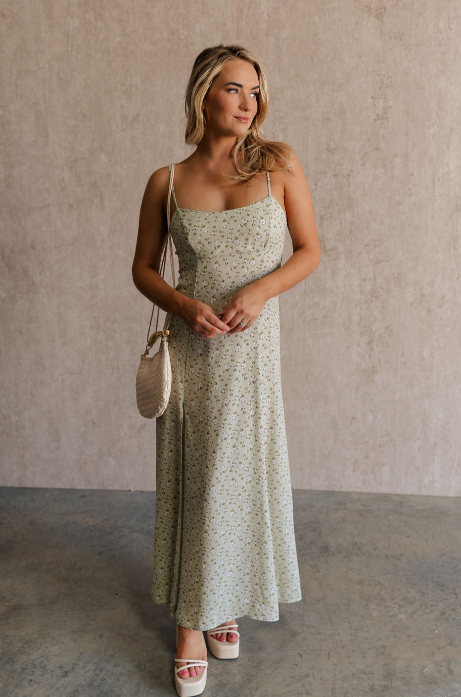 Full body front view of female model wearing the Kaitlyn Sage Floral Maxi Dress that has sage green fabric with a white floral print, thin straps,a side slit and an open back with a tie.