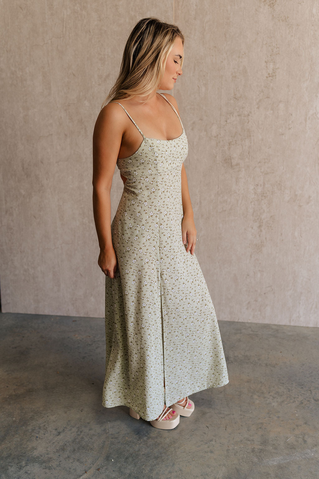 Full body side view of female model wearing the Kaitlyn Sage Floral Maxi Dress that has sage green fabric with a white floral print, thin straps,a side slit and an open back with a tie.