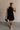 Full body front view of female model wearing the Mara Sleeveless Mock Neck Tie Back Dress in Black that has a ruffle mock neck, mini length hem, and tie back.