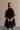 Upper body front view of female model wearing the Mara Sleeveless Mock Neck Tie Back Dress in Black that has a ruffle mock neck, mini length hem, and tie back.