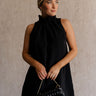 Upper body front view of female model wearing the Mara Sleeveless Mock Neck Tie Back Dress in Black that has a ruffle mock neck, mini length hem, and tie back.