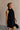 Upper body side view of female model wearing the Mara Sleeveless Mock Neck Tie Back Dress in Black that has a ruffle mock neck, mini length hem, and tie back.