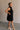 Full body side view of female model wearing the Mara Sleeveless Mock Neck Tie Back Dress in Black that has a ruffle mock neck, mini length hem, and tie back.