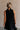 Upper body back view of female model wearing the Mara Sleeveless Mock Neck Tie Back Dress in Black that has a ruffle mock neck, mini length hem, and tie back.