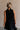 Upper body back view of female model wearing the Mara Sleeveless Mock Neck Tie Back Dress in Black that has a ruffle mock neck, mini length hem, and tie back.