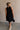 Full body back view of female model wearing the Mara Sleeveless Mock Neck Tie Back Dress in Black that has a ruffle mock neck, mini length hem, and tie back.