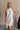 Upper body front view of female model wearing the Mara Sleeveless Mock Neck Tie Back Dress in Off White that has a ruffle mock neck, mini length hem, and tie back.
