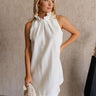 Upper body front view of female model wearing the Mara Sleeveless Mock Neck Tie Back Dress in Off White that has a ruffle mock neck, mini length hem, and tie back.