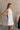 Upper body side view of female model wearing the Mara Sleeveless Mock Neck Tie Back Dress in Off White that has a ruffle mock neck, mini length hem, and tie back.