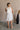 Full body back view of female model wearing the Mara Sleeveless Mock Neck Tie Back Dress in Off White that has a ruffle mock neck, mini length hem, and tie back.