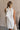 Upper body back view of female model wearing the Mara Sleeveless Mock Neck Tie Back Dress in Off  White that has a ruffle mock neck, mini length hem, and tie back.