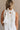 Upper body back view of female model wearing the Mara Sleeveless Mock Neck Tie Back Dress in Off White that has a ruffle mock neck, mini length hem, and tie back.