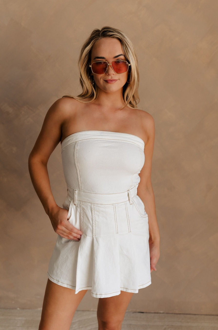 Upper body front view of female model wearing the Kelsey White Denim Romper that has white denim fabric, a strapless neck, and a pleated skirt over shorts lining.