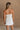 Upper body back view of female model wearing the Kelsey White Denim Romper that has white denim fabric, a strapless neck, and a pleated skirt over shorts lining.