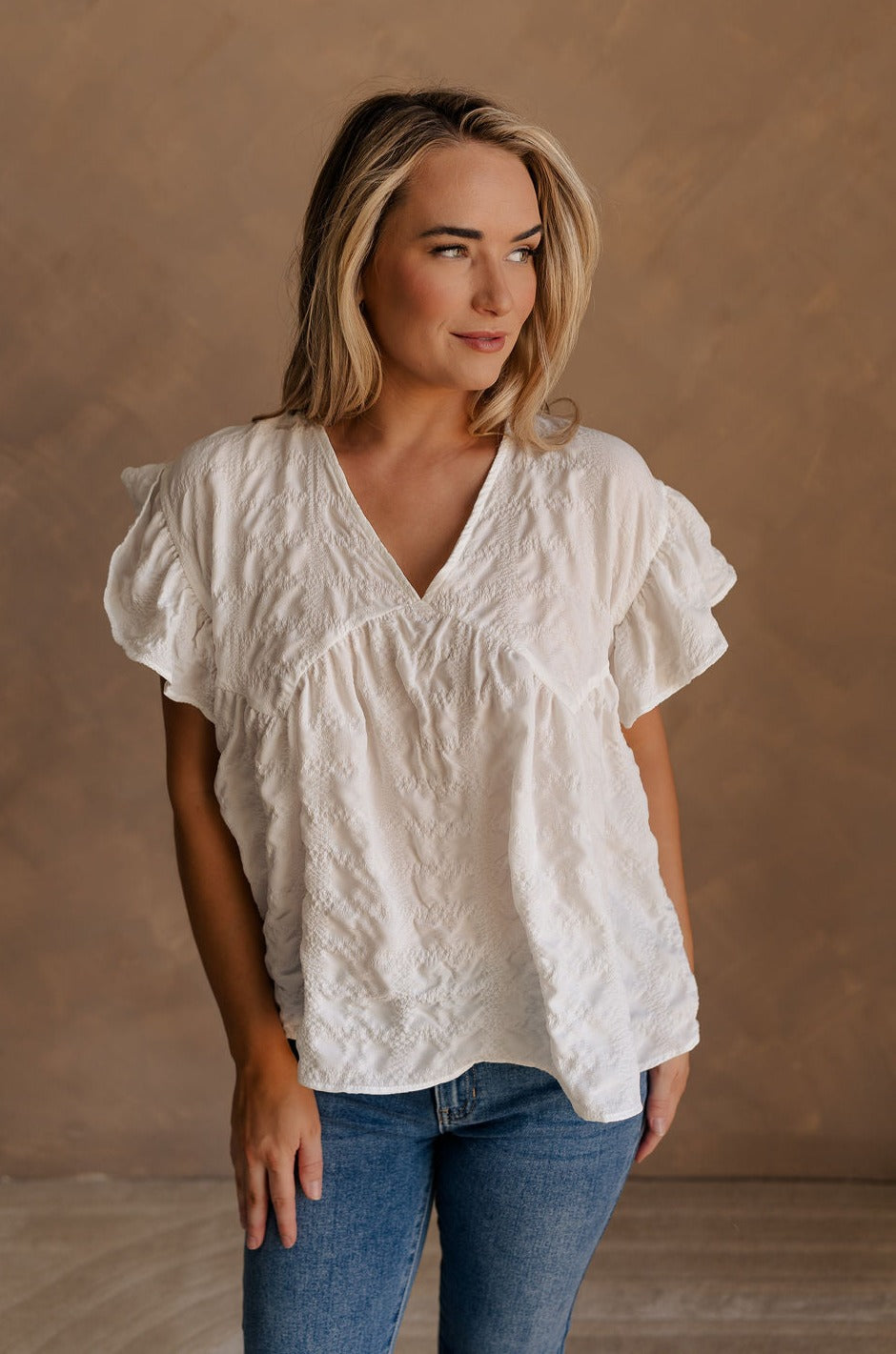 Upper body front view of female model wearing the Vivienne White Textured Top that has white textured fabric, short ruffled sleeves, and v neckline. worn with jeans.