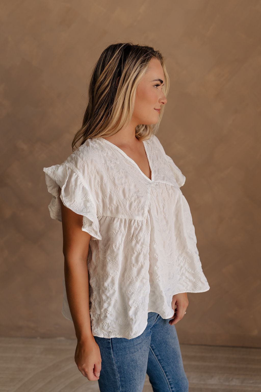 Upper body side view of female model wearing the Vivienne White Textured Top that has white textured fabric, short ruffled sleeves, and v neckline. worn with jeans.