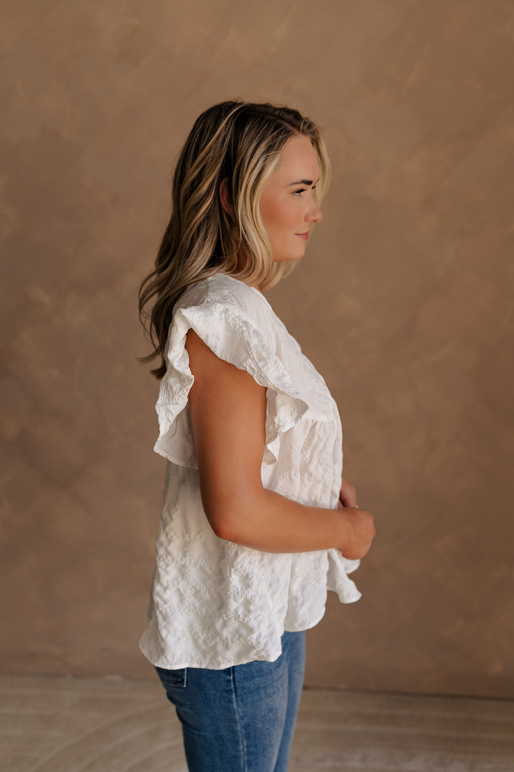 Upper body side view of female model wearing the Vivienne White Textured Top that has white textured fabric, short ruffled sleeves, and v neckline. worn with jeans.