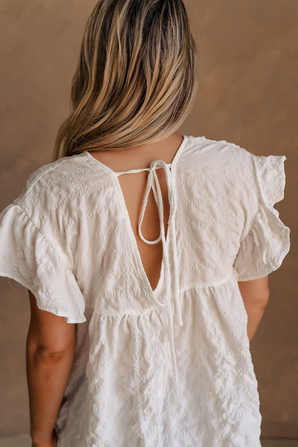 Upper body back view of female model wearing the Vivienne White Textured Top that has white textured fabric, short ruffled sleeves, and v neckline. worn with jeans.