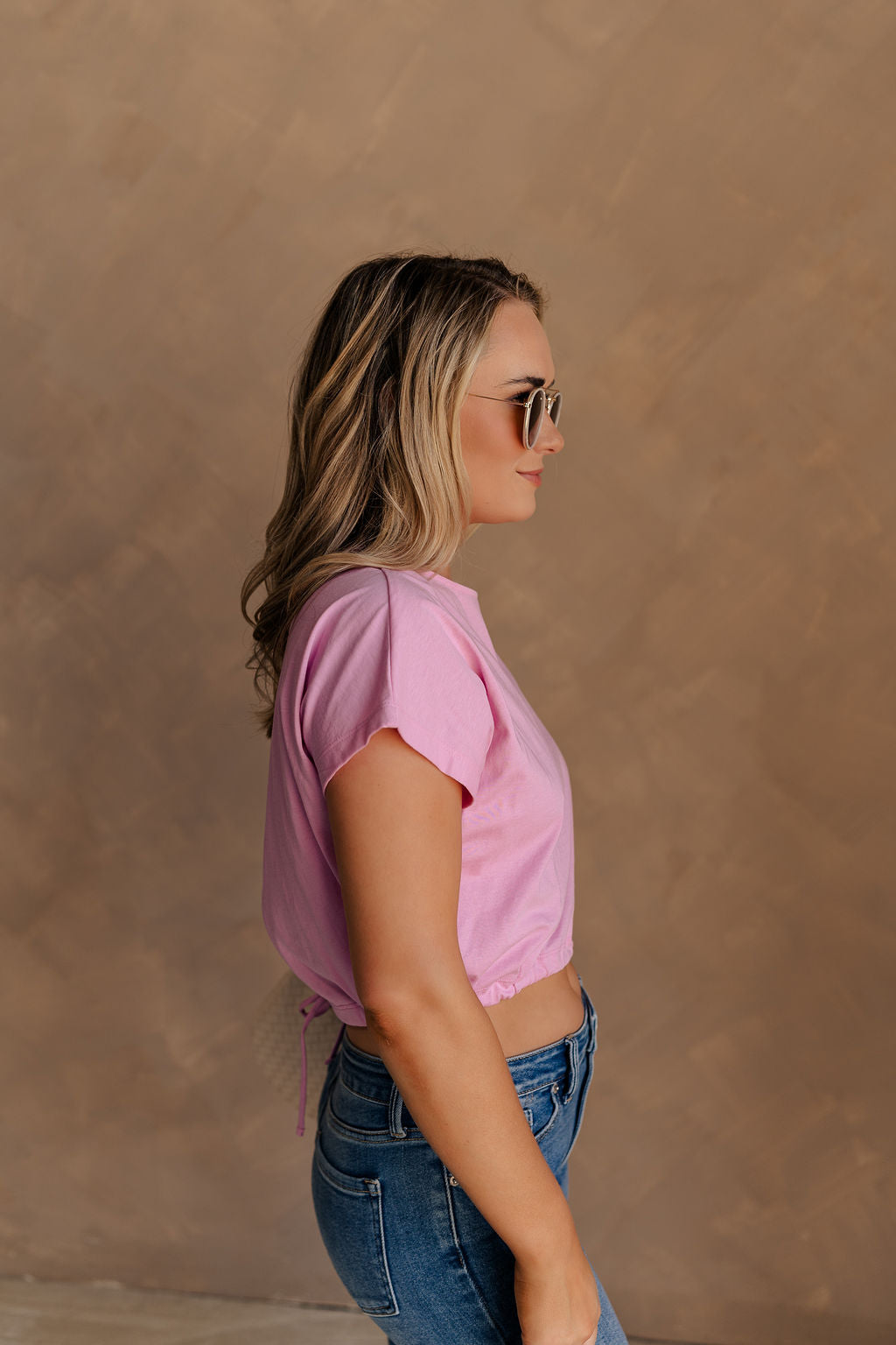 Upper body side view of female model wearing the Belle Pink Short Sleeve Crop Top that has pink fabric, short sleeves, a round neck, and a cropped waist. Worn with jeans.
