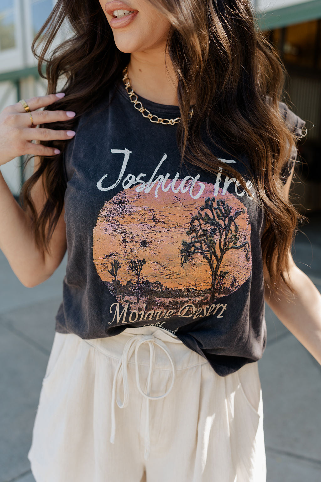 Close front upper body view of female model wearing the Joshua Tree Charcoal Grey Graphic Tee that has distressed charcoal grey fabric, a round neck, and is sleeveless. Features a circular sunset desert graphic with text that says "Joshua Tree Mojave Desert California"