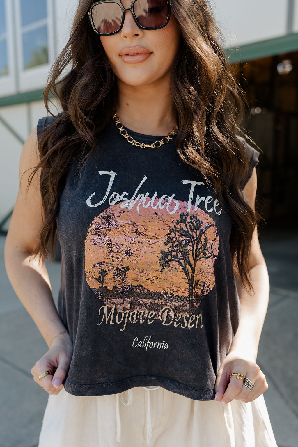 Front upper body view of female model wearing the Joshua Tree Charcoal Grey Graphic Tee that has distressed charcoal grey fabric, a round neck, and is sleeveless. Features a circular sunset desert graphic with text that says "Joshua Tree Mojave Desert California"