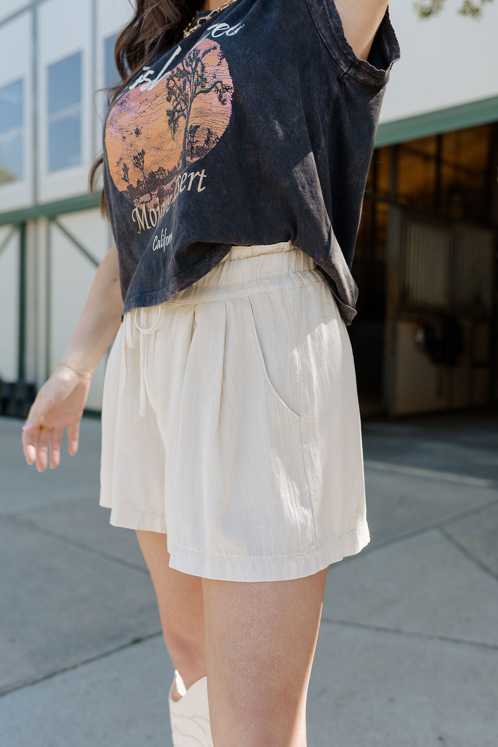 Close=up side view of female model wearing the Jayla Beige Elastic Waist Shorts that have beige lightweight fabric, side pockets, and an elastic waist with tie. Worn with grey tshirt.