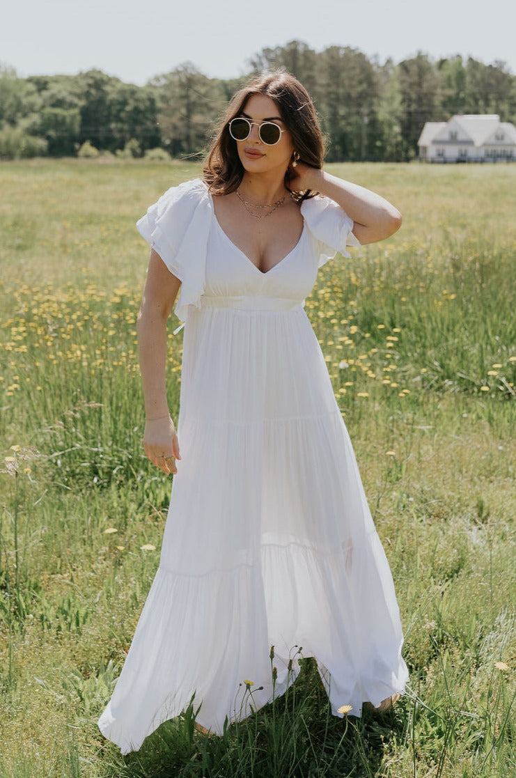 Full body view of female model wearing the Leila Cream Ruffle Short Sleeve Maxi Dress which features White Rayon Fabric, White Lining, Maxi Length, tiered Body, V-Neckline with Ruffle Short Sleeves and Open Back with Back Tie Detail