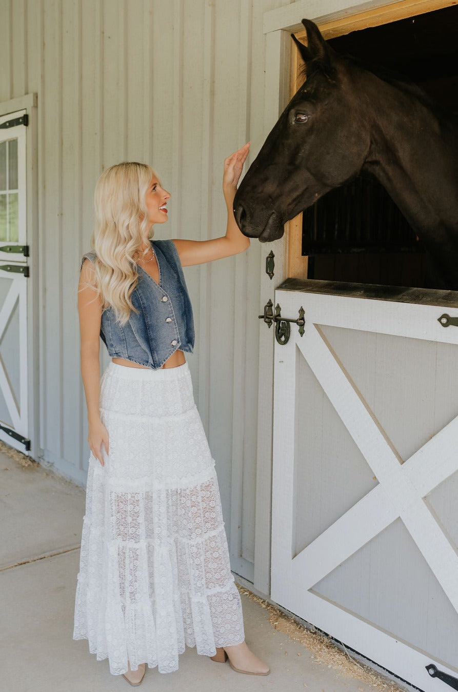 Full body front view of female model wearing the Shiloh White Lace Maxi Skirt that has white lace fabric, half lining, and an elastic waist. model shown petting horse.