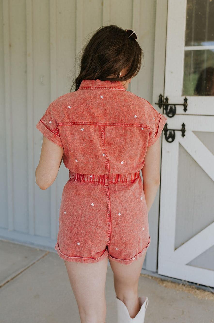 Back view of female model wearing the Krystal Washed Red Denim Star Romper that has red denim fabric with white stars, a snap up front, collar, belt loops, pocket, and short sleeves.