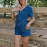 Front view of female model wearing the Mavis Denim Short Sleeve Zip-Up Romper in Medium Wash which features Denim Fabric, Two Front Pockets, Front Zipper Closure, Adjustable Monochrome Belt with Loops, Collared Neckline and Short Sleeves