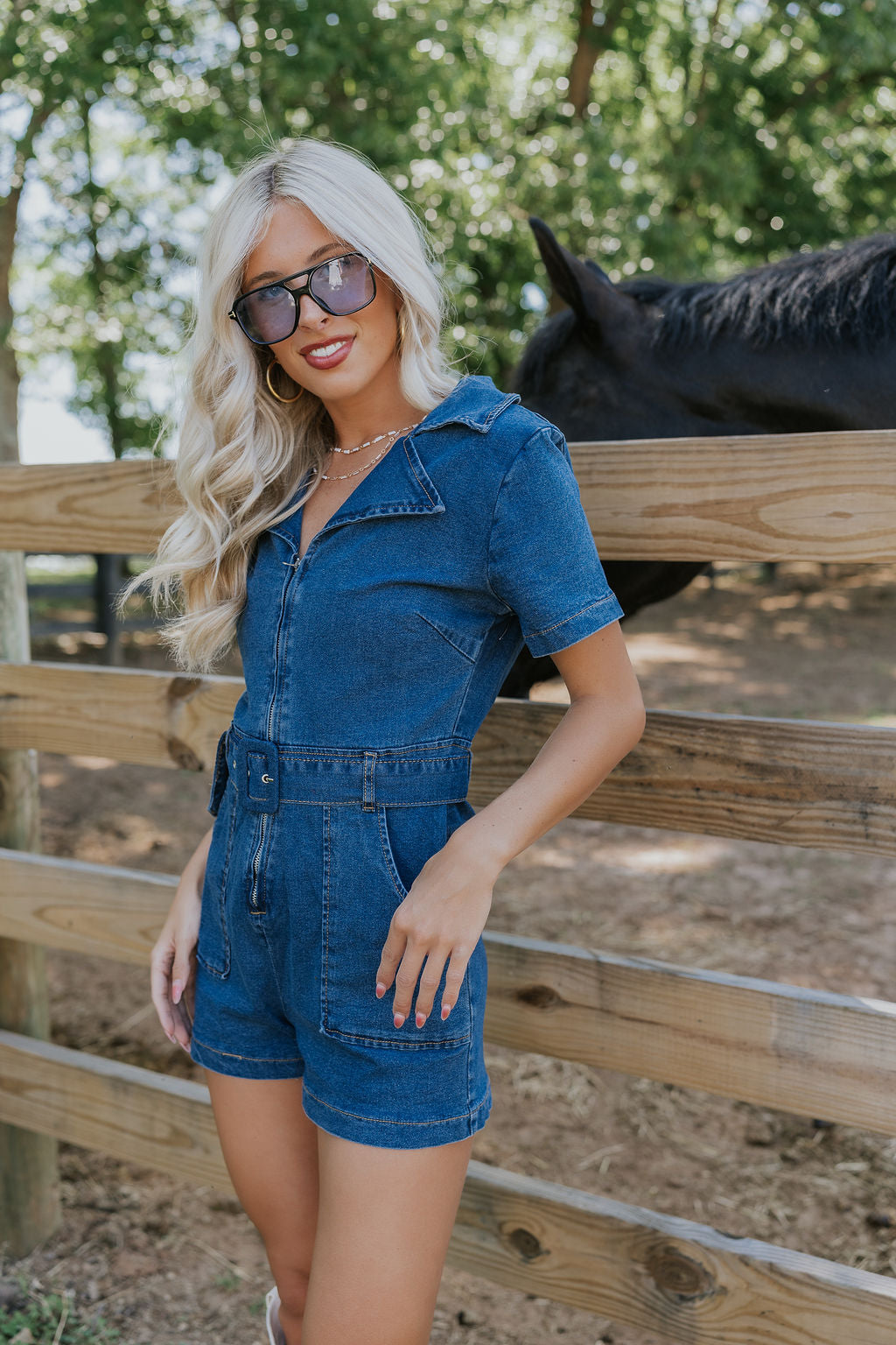 Front view of female model wearing the Mavis Denim Short Sleeve Zip-Up Romper in Medium Wash which features Denim Fabric, Two Front Pockets, Front Zipper Closure, Adjustable Monochrome Belt with Loops, Collared Neckline and Short Sleeves