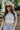 Upper body front view of brunette model wearing the Kira Short Sleeve Cropped Top in ivory that has ivory fabric, short sleeves, a round neck, and cropped waist. Worn with jeans and belt.