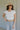 Upper body front view of brunette model wearing the Kira Short Sleeve Cropped Top in ivory that has ivory fabric, short sleeves, a round neck, and cropped waist. Worn with jeans and belt.