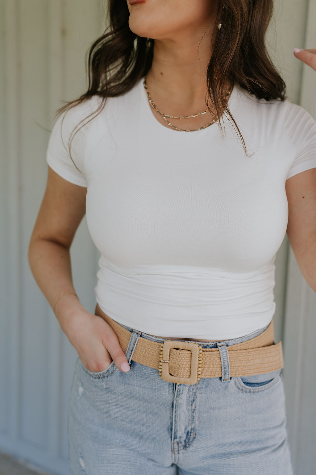 Close up upper body front view of brunette model wearing the Kira Short Sleeve Cropped Top in ivory that has ivory fabric, short sleeves, a round neck, and cropped waist. Worn with jeans and belt.