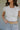 Close up upper body front view of brunette model wearing the Kira Short Sleeve Cropped Top in ivory that has ivory fabric, short sleeves, a round neck, and cropped waist. Worn with jeans and belt.