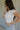 Upper body side view of brunette model wearing the Kira Short Sleeve Cropped Top in ivory that has ivory fabric, short sleeves, a round neck, and cropped waist. Worn with jeans and belt.