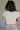 Upper body back view of brunette model wearing the Kira Short Sleeve Cropped Top in ivory that has ivory fabric, short sleeves, a round neck, and cropped waist. Worn with jeans and belt.